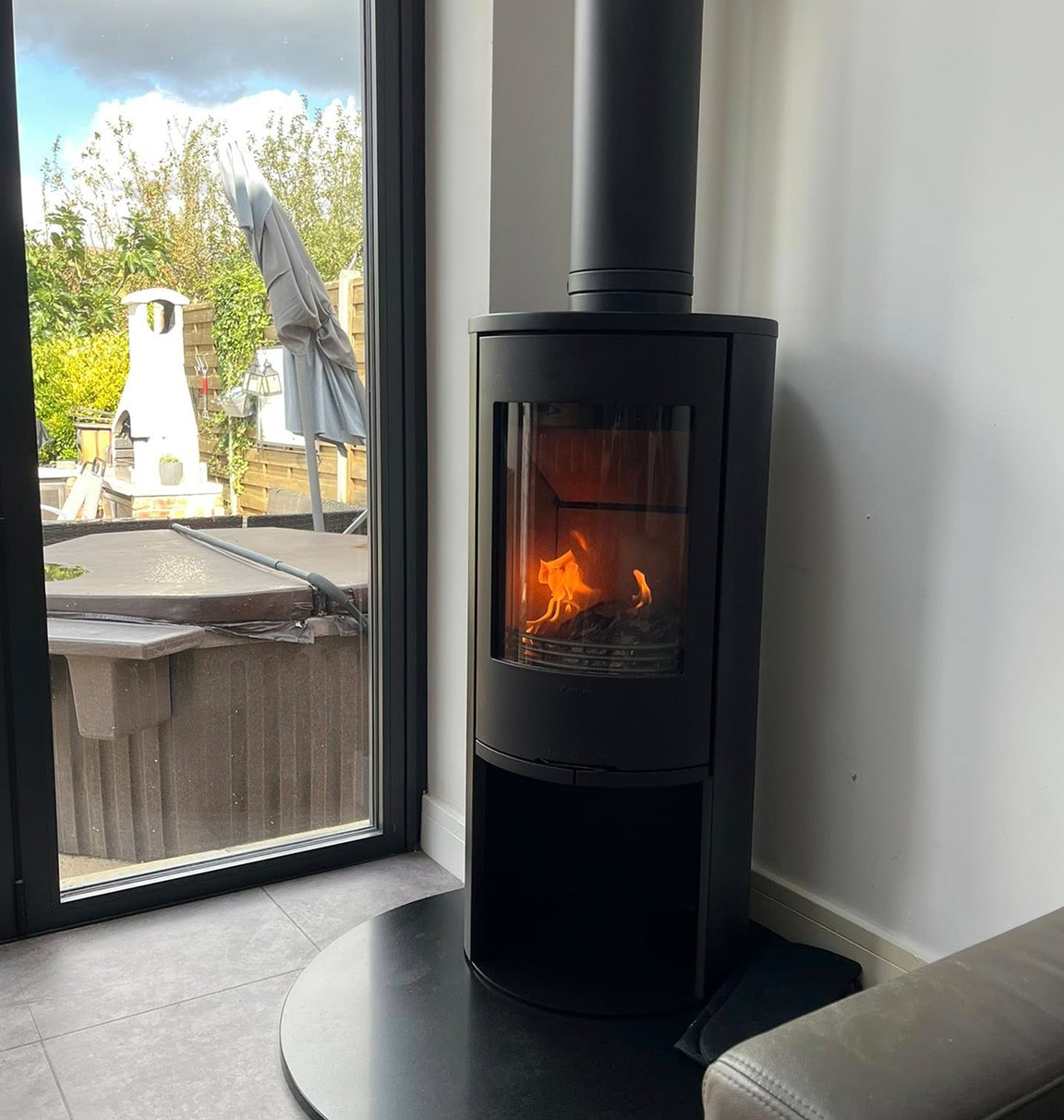 Cylindrical wood burning stove in the corner of a grey room by a window overlooking a hot tub in the garden