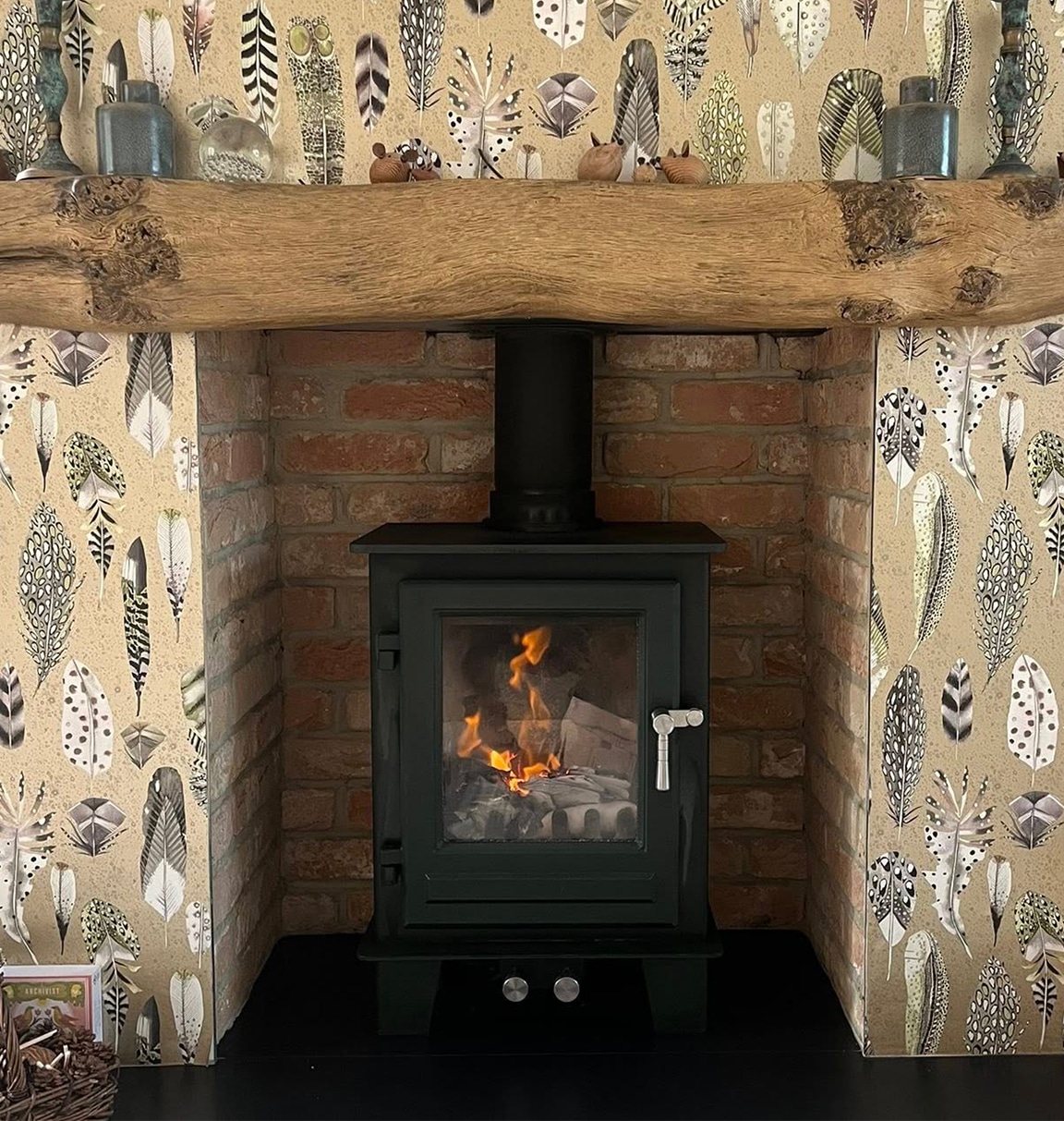 Client photo of a black square wood burner in an exposed brick fireplace with feather wallpaper and a wooden floating mantel